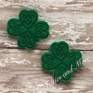 以下が含まれることがあります： ケルト結び目のデザインが施された緑色のフェルト製のシャムロックパッチが2つ。シャムロックは、「Wet Grass Felties and More」というテキストが書かれた木製の背景の上に置かれています。