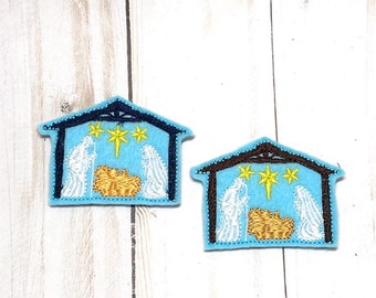 May include: Two felt Christmas decorations depicting the nativity scene. The decorations are blue with a yellow star and white figures. One decoration has a dark blue stable and the other has a brown stable.