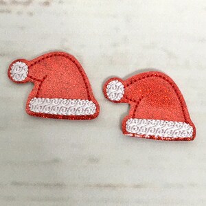 May include: Two red glitter Santa hats with white trim. The hats are on a white wooden background.