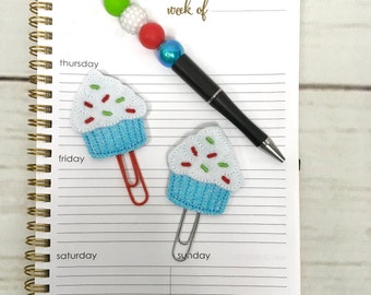 May include: A white notebook with gold spiral binding and lined pages. The notebook has the words "week of" written on the top. There are two blue and white cupcake-shaped paper clips with red and green sprinkles on the notebook. The notebook also has the words "thursday", "friday", "saturday", and "sunday" written on it.