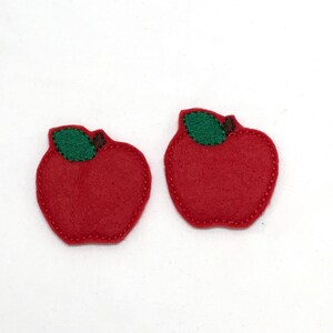 Peut inclure: Deux formes de pommes en feutre rouge avec des feuilles en feutre vert et des tiges brunes. Les pommes sont cousues avec du fil rouge.