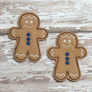 May include: Two brown felt gingerbread people with white icing and blue buttons. The gingerbread people are on a white wooden background.