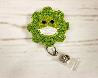 Könnte beinhalten: Grüner Glitzer-Badgehalter mit einem grünen und weißen Gesichtsmasken-Design. Die Gesichtsmaske hat zwei schwarze Augen. Der Badgehalter hat eine transparente, einziehbare Schnur.