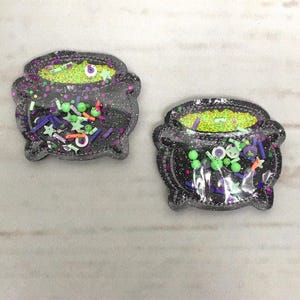 Pode incluir: Dois patches de ferro em forma de caldeirão preto com glitter, com confeitos verdes, laranja e roxos dentro.