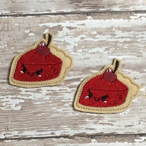 May include: Two red pie slice patches with a cherry on top and a smiling face, outlined in tan and stitched with black thread. The patches are on a distressed, white-painted wooden surface.