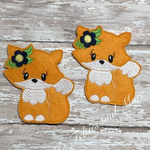 Puede incluir: Dos apliques de zorro de fieltro naranja con caras blancas y ojos negros. Cada zorro tiene una flor verde y amarilla con un centro azul en la cabeza. Los zorros están sobre un fondo de madera blanco.