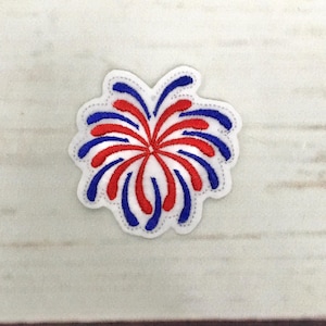 Puede incluir: Un parche bordado blanco con forma de fuegos artificiales. El diseño presenta líneas curvas rojas y azules que irradian desde un punto central. El parche tiene un borde blanco con una puntada punteada.