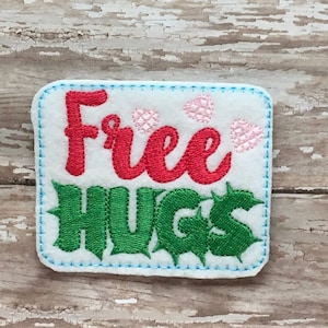 以下が含まれることがあります： 縁が青いステッチで縫われた白いフェルトのパッチ。パッチには「Free Hugs」という文字が赤と緑で刺繍されています。