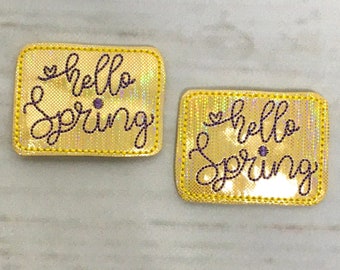 Puede incluir: Dos parches termoadhesivos de color dorado con el texto "hello spring" en letras cursivas moradas. Los parches tienen un acabado brillante.