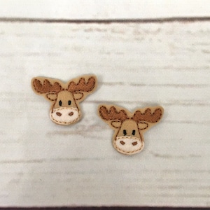 以下が含まれることがあります： 刺繍のディテールが施された、茶色のフェルト製のムースのアップリケが2つ。ムースは茶色の目、茶色の鼻、白い口吻をしています。
