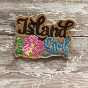 May include: Embroidered patch with the text "Island Girl" in brown and blue lettering. A pink hibiscus flower with green leaves is to the left of the text. The patch is on a light brown wooden surface.