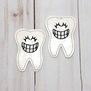 May include: Two white felt tooth shapes with black embroidered smiling faces. The teeth have closed eyes and a row of teeth showing.