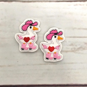以下が含まれることがあります： ピンクのテンガロンハットとピンクのブーツを履いたガチョウの白いフェルトパッチが2つ。それぞれのガチョウは赤いハートを持っています。