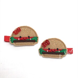 May include: Two felt hamburger hair clips with red bows and green lettuce. The hamburgers have a smiling face and a brown patty.
