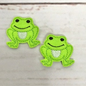 Könnte beinhalten: Zwei grüne Filz-Frosch-Applikationen mit schwarzen Augen und einem weißen Lächeln. Die Frösche sind mit grünem Garn gestickt.