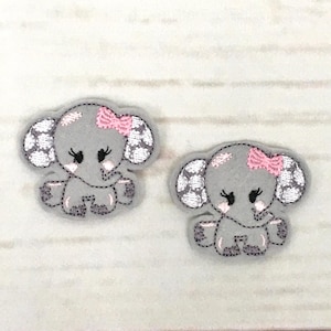 以下が含まれることがあります： ピンクのドット、ピンクのリボン、まつげが付いた、漫画の象のグレーのフェルトアップリケ2つ。