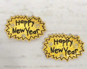 Puede incluir: Dos parches de planchado de purpurina dorada con el texto "Happy New Year" en negro. Los parches tienen forma de estrella.