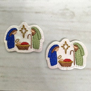 May include: Two embroidered felt patches depicting the nativity scene. Each patch features a blue and green figure, a baby in a manger, a star, and a shepherd's staff.