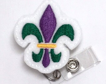 May include: White felt badge holder with a purple and green embroidered fleur-de-lis design. The badge holder has a clear plastic clip.