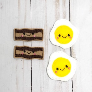 May include: Two felt patches, one with a brown and white bacon design and the other with a white and yellow fried egg design. Both patches have a smiling face with pink cheeks.