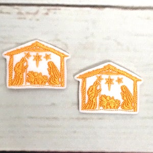 May include: Two embroidered patches depicting a gold Nativity scene within a white house-shaped frame. The scene shows Mary, Joseph, and baby Jesus, with stars above. The patches are on a light wood-grain surface.