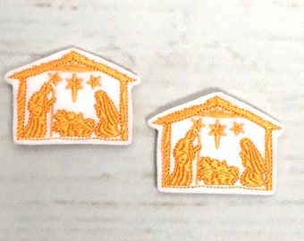 May include: Two embroidered patches depicting a gold Nativity scene within a white house-shaped frame. The scene shows Mary, Joseph, and baby Jesus, with stars above. The patches are on a light wood-grain surface.