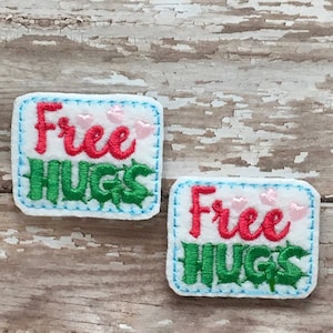 以下が含まれることがあります： 青いステッチが縁取りされた白いフェルト生地の2つのパッチ。各パッチには、赤い文字と緑の文字で「Free Hugs」という文字が書かれており、その上にピンクのハートが描かれています。