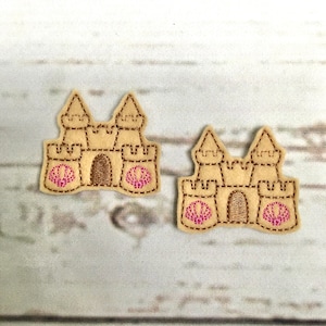 以下が含まれることがあります： ピンクの貝殻のディテールが付いた、茶色のフェルト製の砂城のアップリケが2つ。砂城は茶色の糸で縫われており、表面にテクスチャがあります。