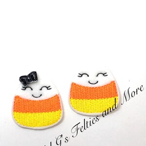 Może przedstawiać: Dwa aplikacje z filcu w kształcie cukierków kukurydzianych z uśmiechającymi się twarzami. Jeden cukierek kukurydziany ma czarną błyszczącą kokardkę na górze.  "Mel G's Felties and More" jest napisane czarnym tekstem na białym tle.