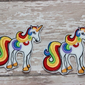 Puede incluir: Dos parches de unicornio de fieltro blanco con crines y colas de arcoíris. Los unicornios están de pie sobre un fondo de madera clara.