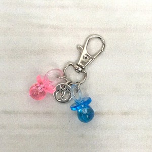 May include: A silver keychain with a silver charm featuring two footprints. Attached to the charm are two pacifiers, one pink and one blue.