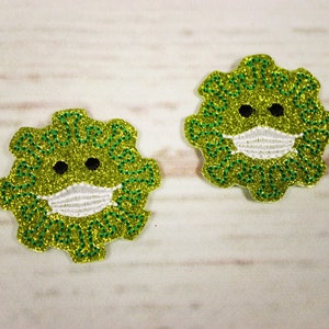 May include: Two green glitter iron-on patches shaped like a virus with a white face mask and black eyes. The text "Stay Safe" is written around the edge of each patch.