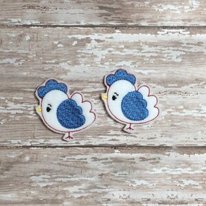 Peut inclure: Deux poussins brodés en feutre blanc avec des ailes bleues et des pieds rouges. Les poussins sont tournés dans des directions différentes.