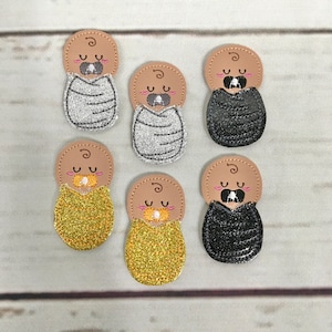 Puede incluir: Seis apliques bordados de bebés. Los bebés tienen piel morena, ojos cerrados y chupetes. Las envolturas son plateadas, negras y doradas con purpurina. Los apliques están dispuestos sobre una superficie de madera blanca.