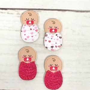 Puede incluir: Cuatro apliques de bebé de fieltro con un chupete en la boca. Dos llevan tela estampada con corazones rosas y blancos y dos llevan tela de purpurina roja.