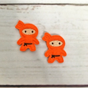 May include: Two orange felt appliques of ninjas with black belts. The ninjas have black eyes and mouths.