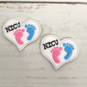 Puede incluir: Dos parches blancos en forma de corazón con el texto "NICU" en negro. Cada parche presenta un diseño de huella rosa y azul. Los parches están cosidos con hilo gris y están sobre una superficie de vetas de madera clara.