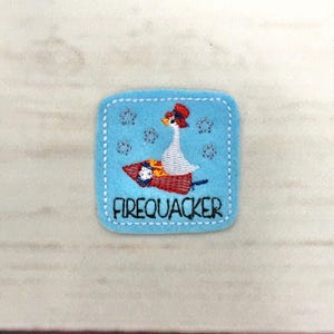 Puede incluir: Un parche cuadrado de fieltro azul claro con un borde cosido blanco. El parche presenta un diseño bordado de un ganso con un sombrero rojo, montado en un cohete. La palabra "FIREQUACKER" está bordada debajo.