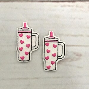 以下が含まれることがあります： ピンクのハートとピンクのストローが付いた白いタンブラーが2つ。タンブラーにはハンドルと蓋が付いています。