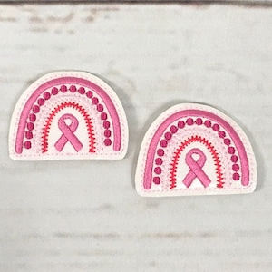 May include: Two pink and red embroidered rainbow patches with a pink ribbon in the center. The patches are on a white wooden background.