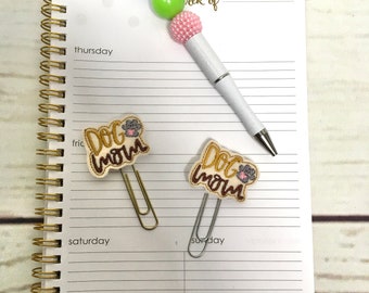 May include: Two gold paper clips with a white fabric embellishment that says "Dog Mom" in brown letters. The paper clips are on a white lined notebook with the words "Thursday", "Friday", "Saturday", and "Sunday" written on the page. A white pen with a pink, green, and blue beaded top is on the notebook.