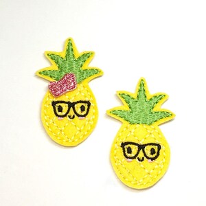May include: Two yellow felt pineapple patches with green tops and black glasses. One pineapple has a pink bow on top.