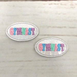 Könnte beinhalten: Zwei weiße, ovale Bügelbilder mit dem Wort "GYMNAST" in Regenbogenfarben. Die Patches haben einen glitzernden Hintergrund.