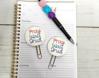 May include: Two white paper clips with a round, white fabric embellishment attached to each. The embellishments have the words "Pray Wait Trust" embroidered on them in red, blue, and purple thread. The paper clips are on a white notebook with gold spiral binding.