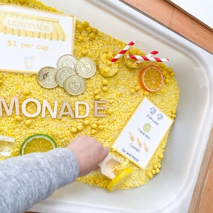 Puede incluir: Una bandeja de plástico blanca llena de arroz amarillo y frijoles amarillos y blancos. Hay letras de madera que deletrean la palabra "MONADE" y un letrero que dice "LEMONADE 1€ por taza". También hay varias monedas de oro, un vaso pequeño con pajitas rojas y blancas, una rodaja de limón y una tarjeta con instrucciones para hacer limonada.