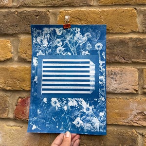 May include: A blue and white floral print artwork with a rectangular section of text. The text is in a rectangular frame with horizontal blue and white stripes. The artwork is clipped to a brick wall with a red clip.