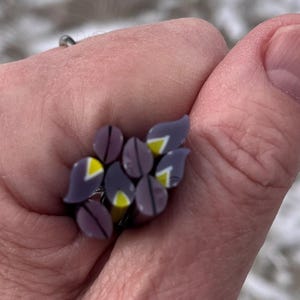 May include: A cluster ring featuring a design of purple and gray glass beads with yellow accents. The ring's design is reminiscent of a flower or a cluster of grapes. The ring is held in a hand.