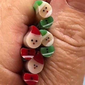 May include: A close-up of several small, decorative Christmas-themed beads. The beads are shaped like Santa Claus and elves, with red, green, white, and beige colors. The beads are held in a hand.
