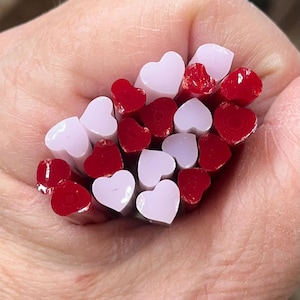 May include: A collection of red and white heart-shaped glass beads. The beads are held in a hand, showcasing their glossy texture and vibrant colors. The hearts are a mix of solid red and opaque white.