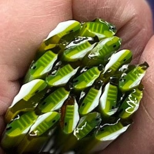 May include: A collection of small, green, and white glass beads, possibly for jewelry making, held in a hand. The beads have a rectangular shape with white accents and a textured appearance.